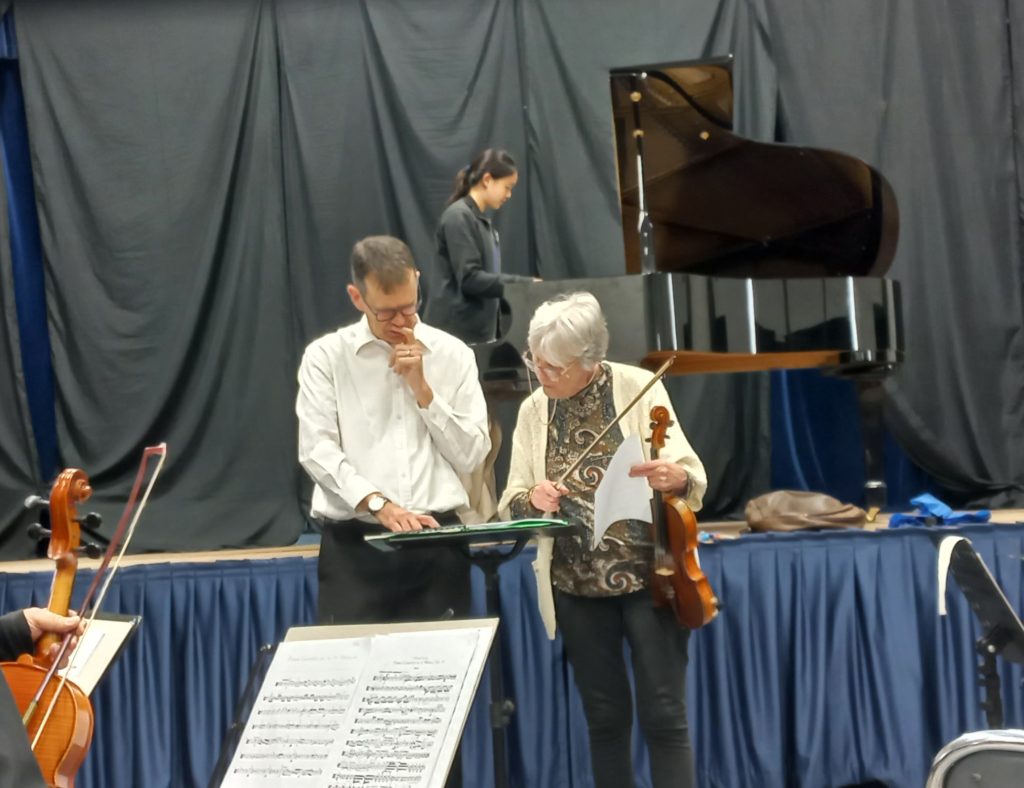 Conductor and Concert Master conferring before a rehearsal of the Grieg piano Concerto's 1st Movement - soloist Ziyu Peng warming up at he piano behind them.